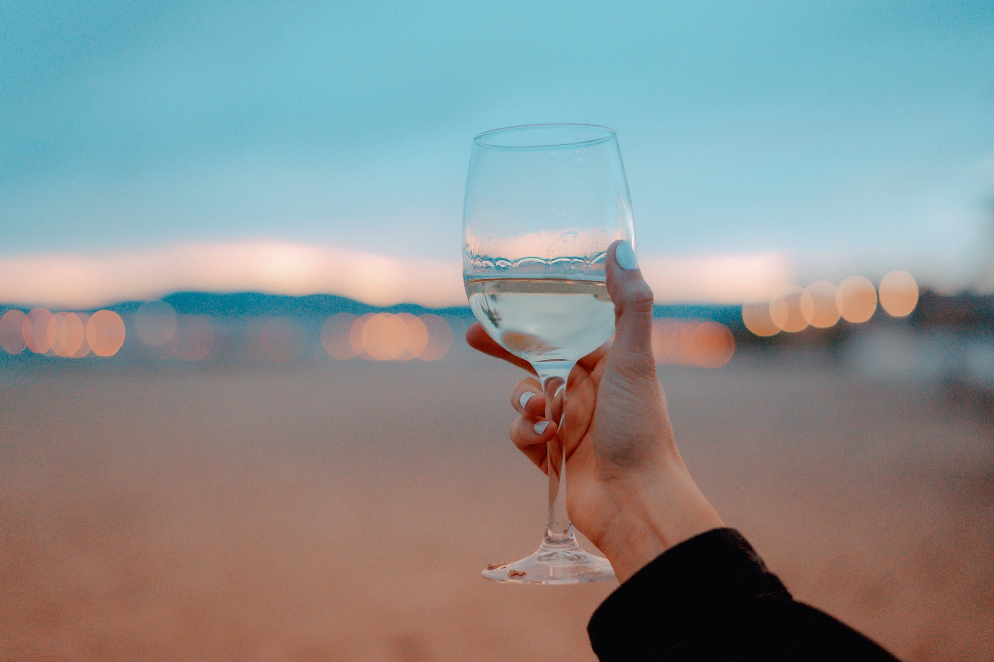 Photo d'un verre de vin blanc tenu par une main de femme devant un paysage d'été au crépuscule - Crédit Photo : Toa Heftiba